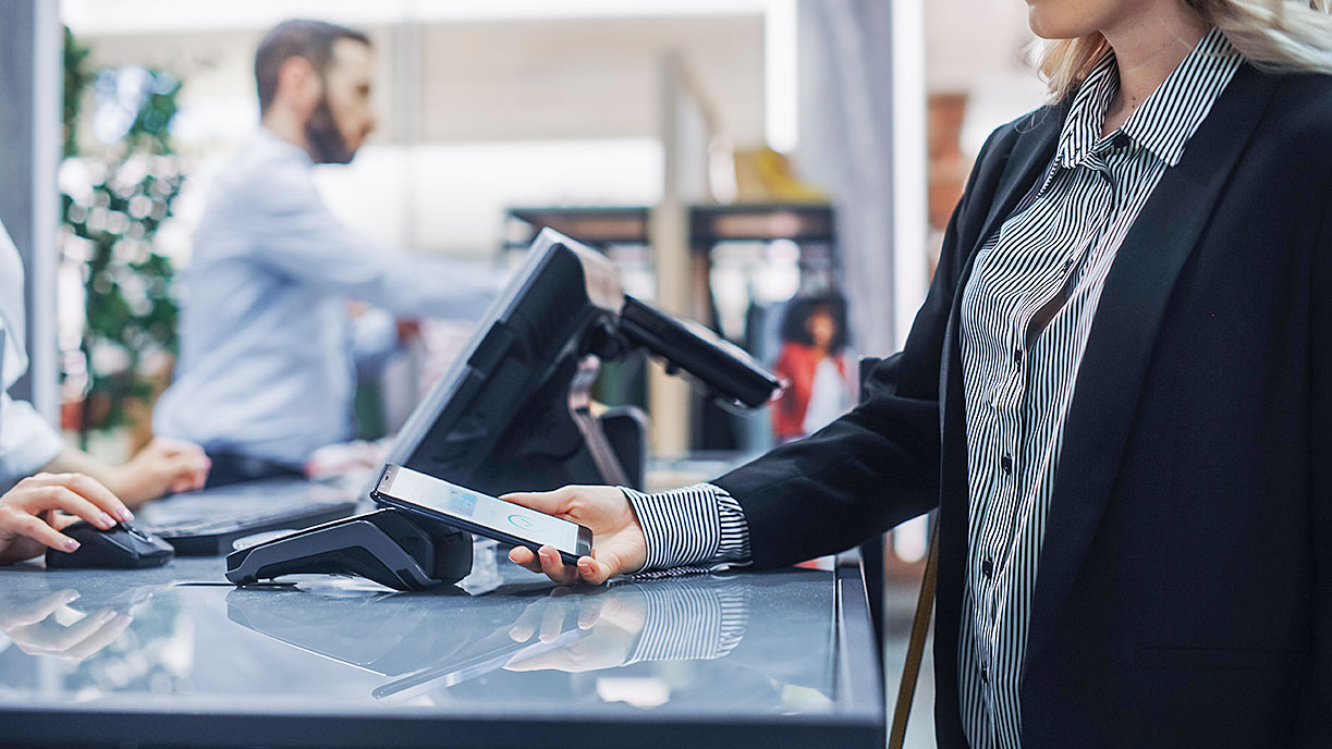 Businesswoman in black blazer holding smartphone near payment terminal on glass counter; male employee assists customer in background.