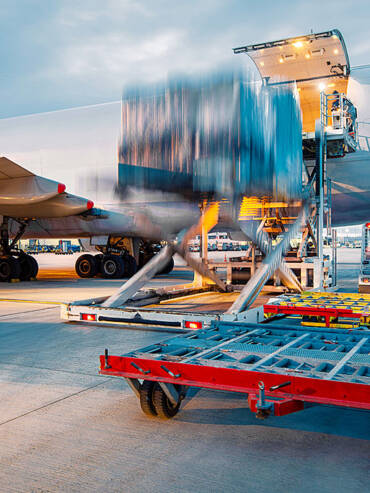 Large cargo airplane with open hold door loading red metal cart on gray tarmac, airport vehicles in background under bright light.
