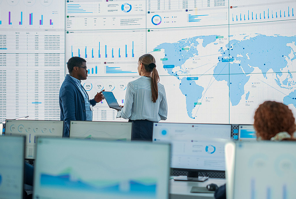 Two diverse professionals analyzing global data visualizations on large wall display in bright computer-filled room.