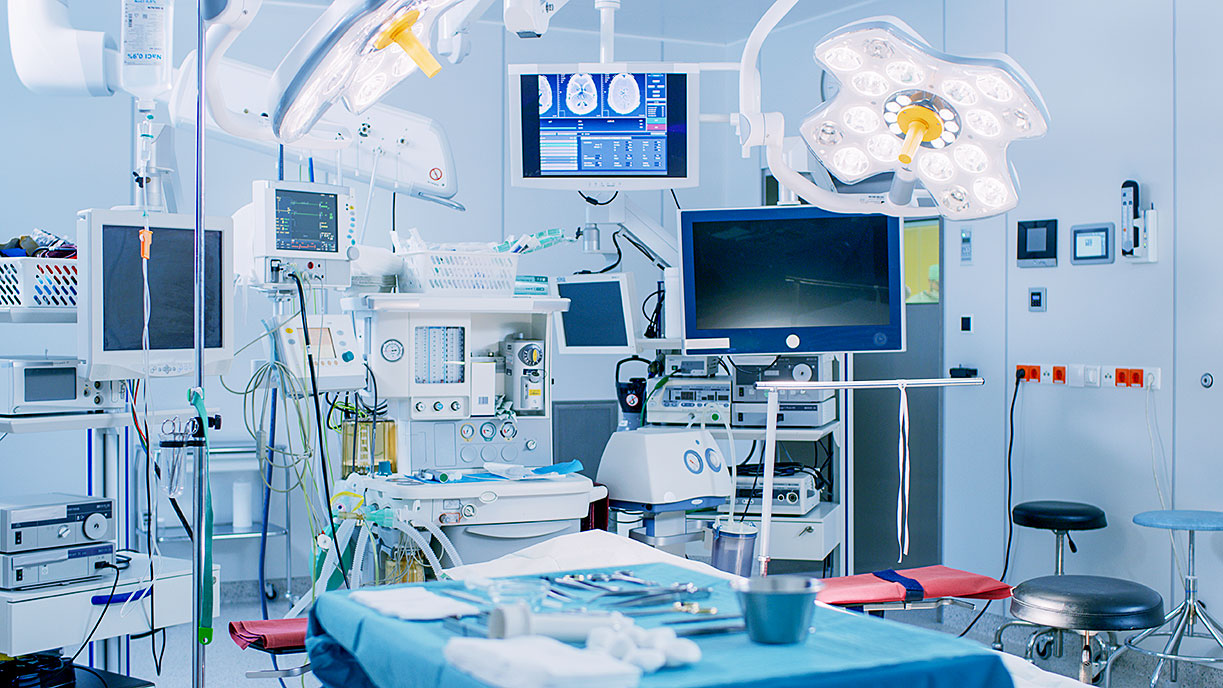 Modern surgical room with operating table, overhead surgical lights, monitors displaying medical data, and organized surgical instruments.
