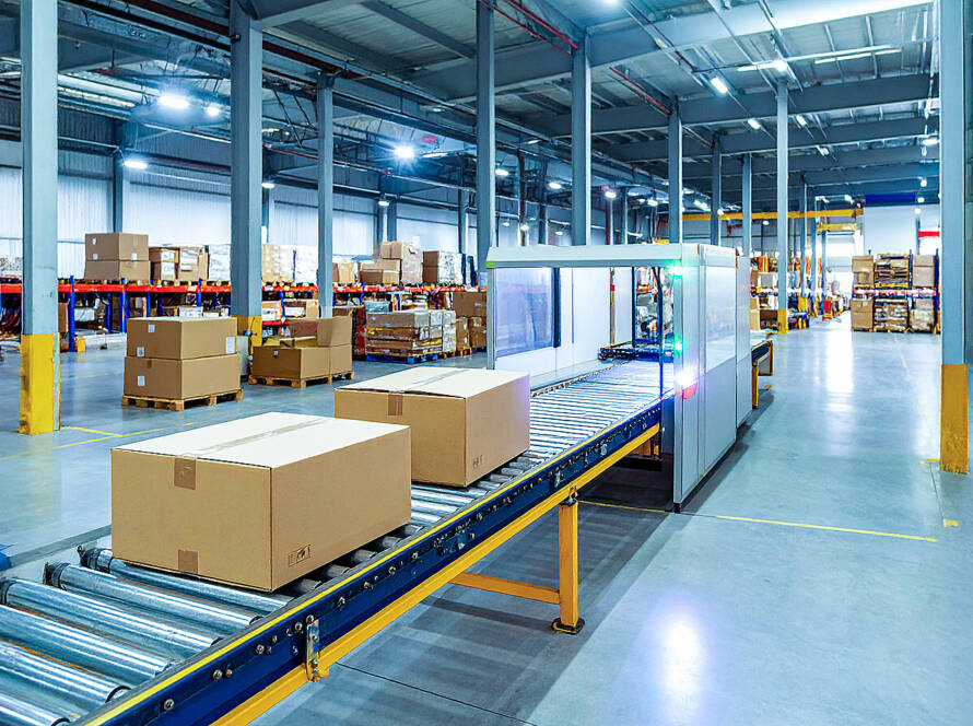High-angle view of conveyor belt in warehouse with brown cardboard boxes, metal supports, transparent enclosure, and green-blue flashing light.