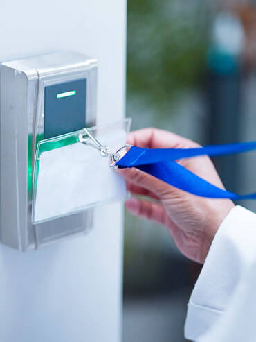 Hand holding security badge with barcode on blue lanyard near card reader and biometric scanner on sleek metallic door frame.