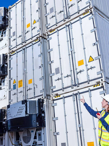 wo women in hard hats and high-visibility vests inspect refrigerated shipping containers with safety labels against clear blue sky.