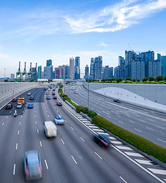 busy-urban-highway-city-skyline-cranes-greenery-singapore Busy urban highway curves with blurred cars; modern city skyline with glass buildings, cranes, and greenery under partly cloudy sky.