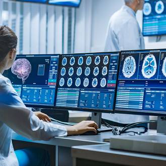 female-scientist-brain-scans-medical-lab-monitors Female scientist in lab coat analyzing brain scans on multiple monitors; two colleagues discuss in background of modern medical lab.